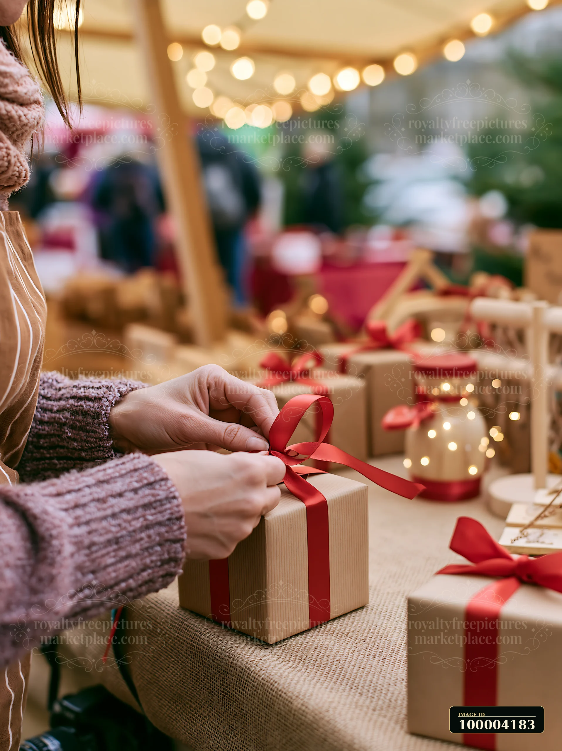 Hands tie a red ribbon around a kraft-wrapped gift box among presents and warm string lights.