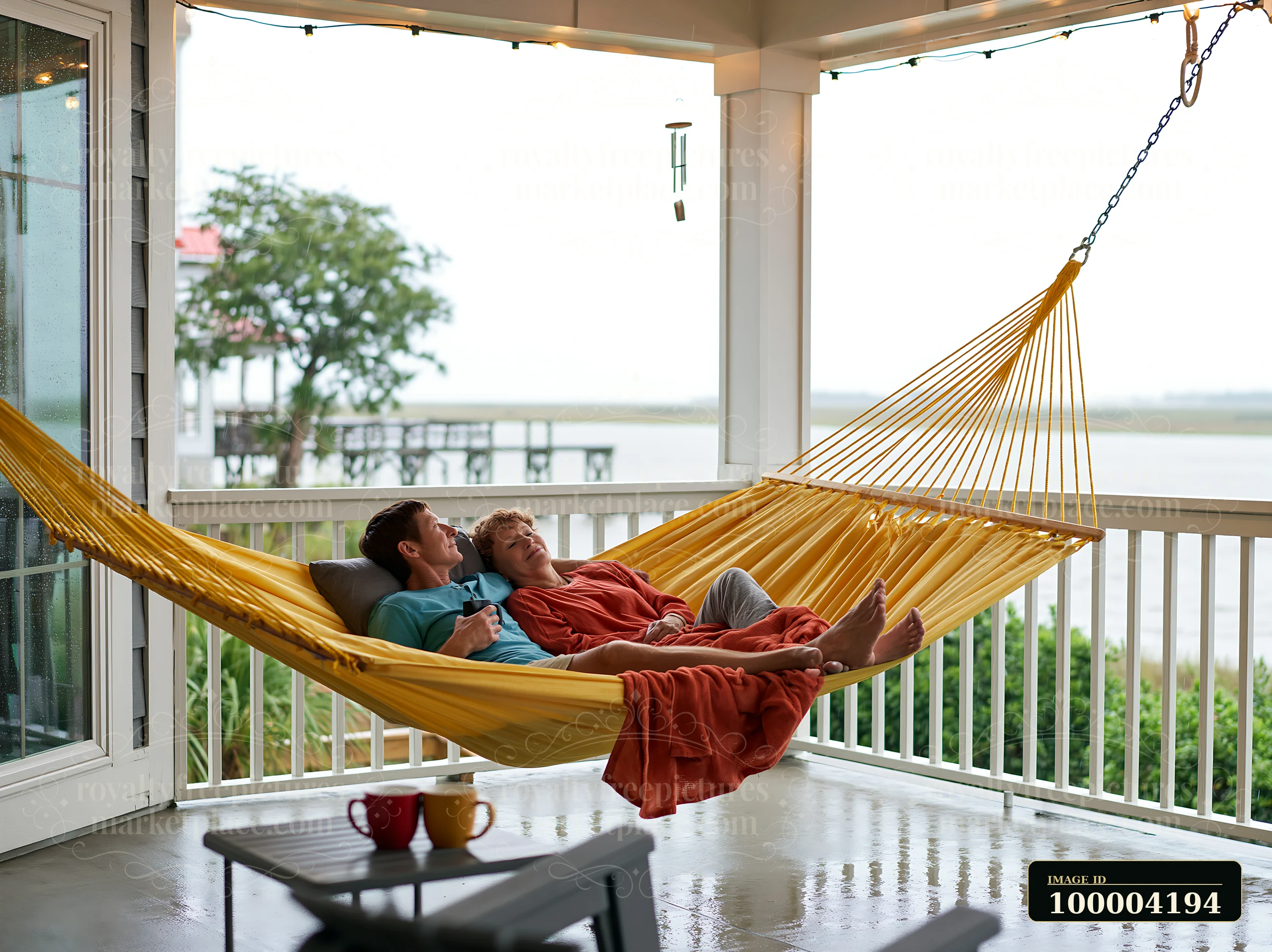 An older couple enjoys their time together in a hammock on a covered porch while it is raining.