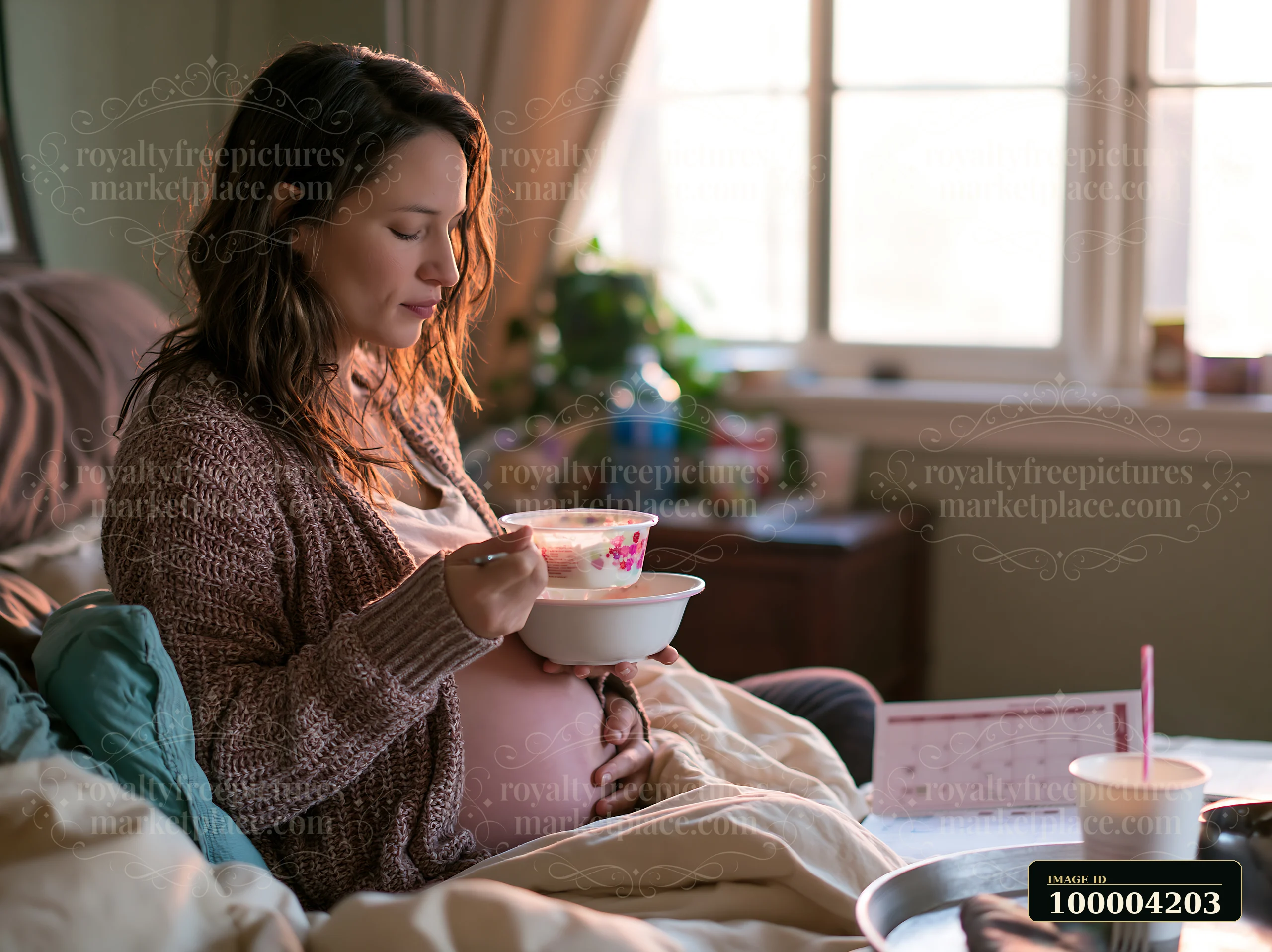 A pregnant woman enjoys breakfast in bed, holding a bowl while seated on a cozy bed.