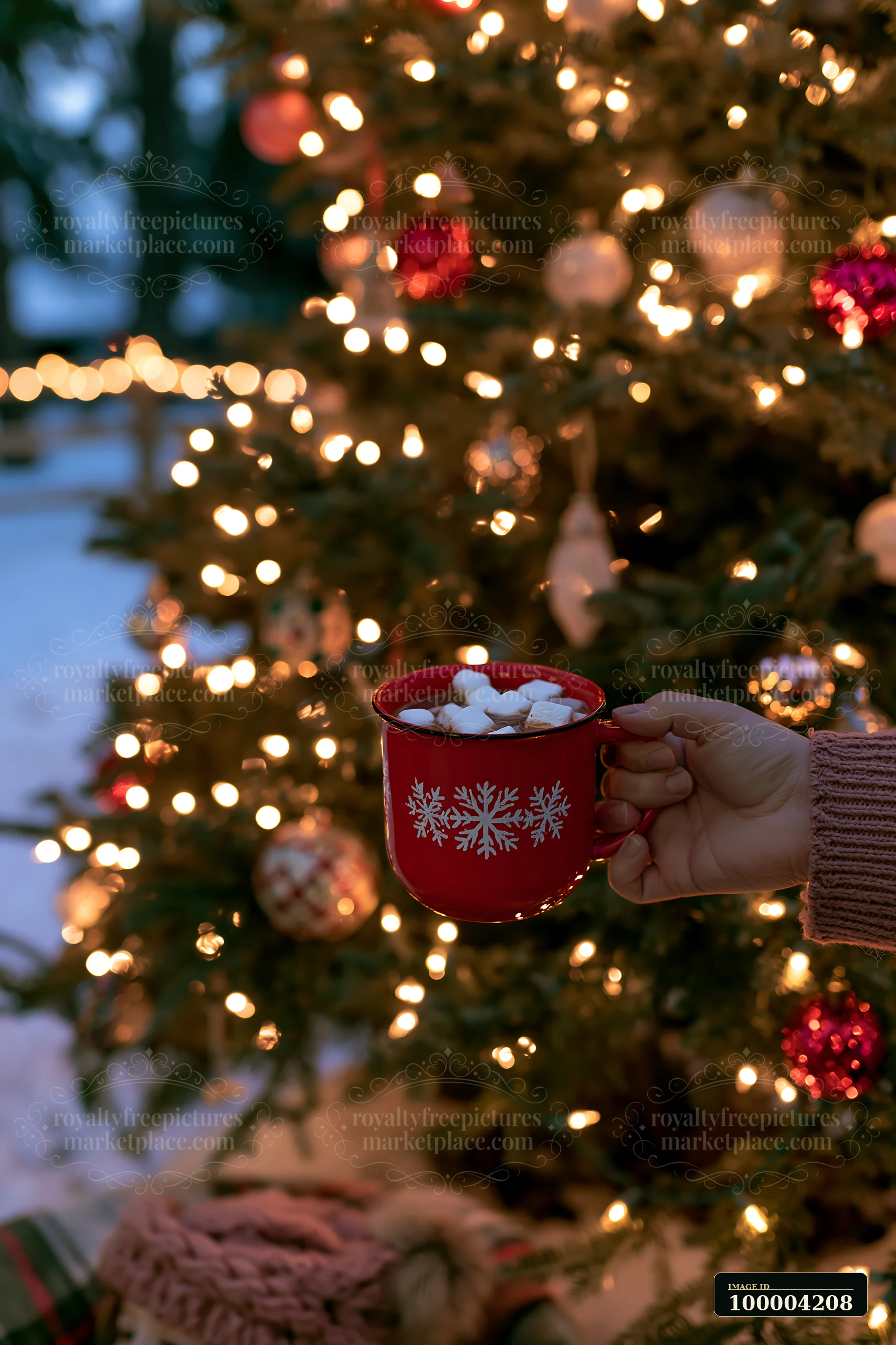 A hand holds a red cup of warm cocoa topped with marshmallows near a decorated tree with lights.