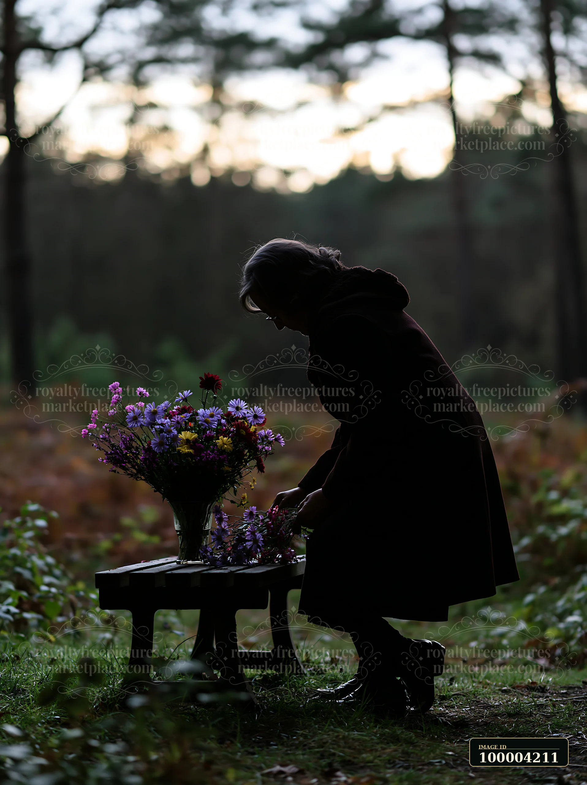 Silhouetted person places flowers on a memorial bench in a dim forest at dusk.