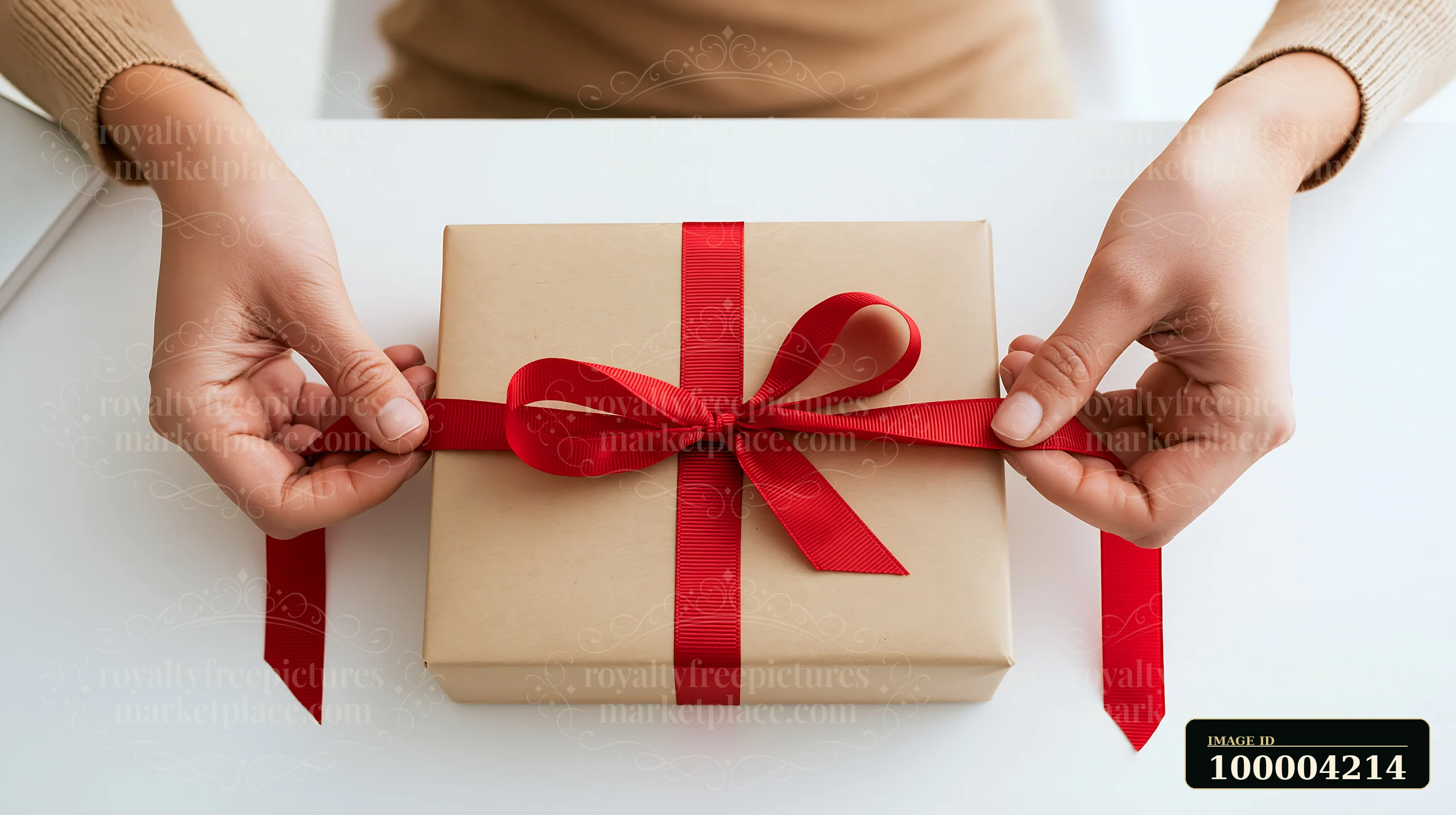Hands tie a red ribbon around a beige wrapped gift box on a white tabletop.