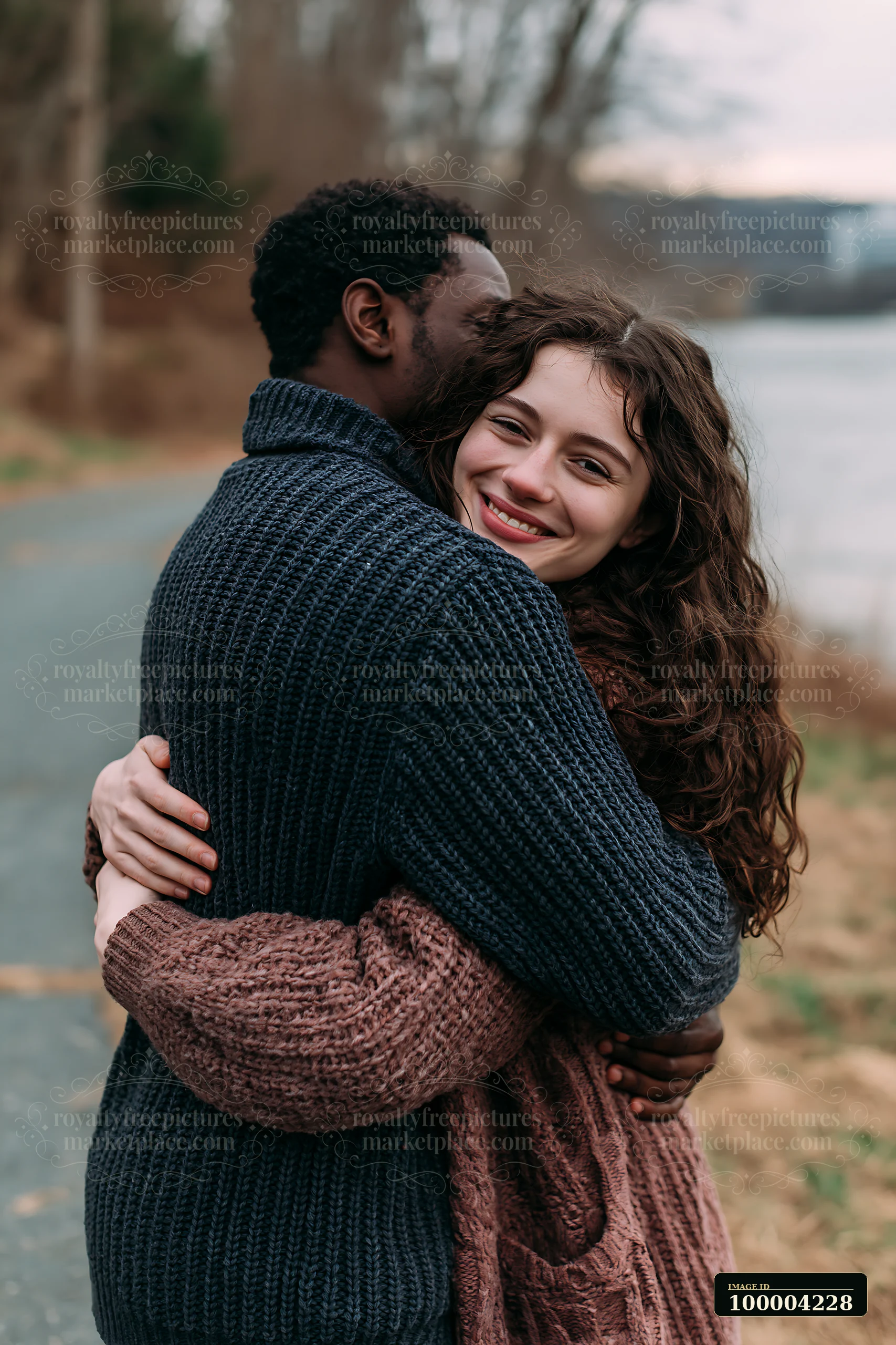 Smiling couple embraces beside a lake outdoors, with blurred trees and calm water in the background.
