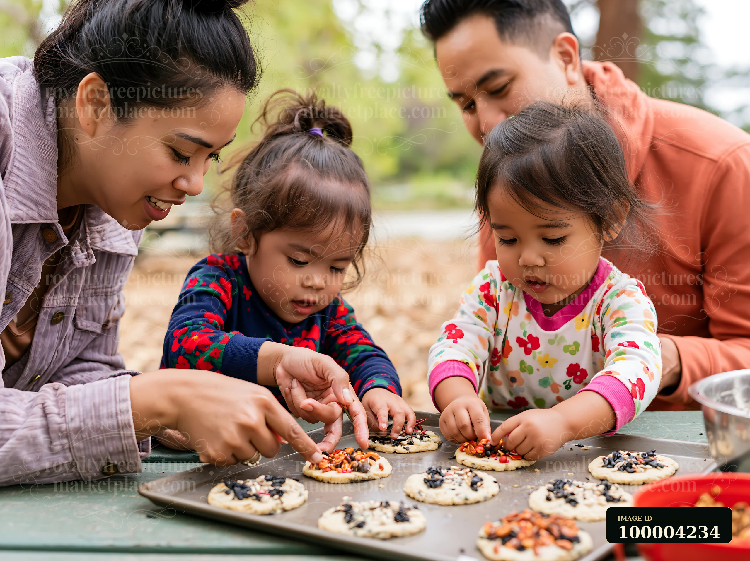Two parents help two children decorate cookies with colorful toppings at an outdoor table.
