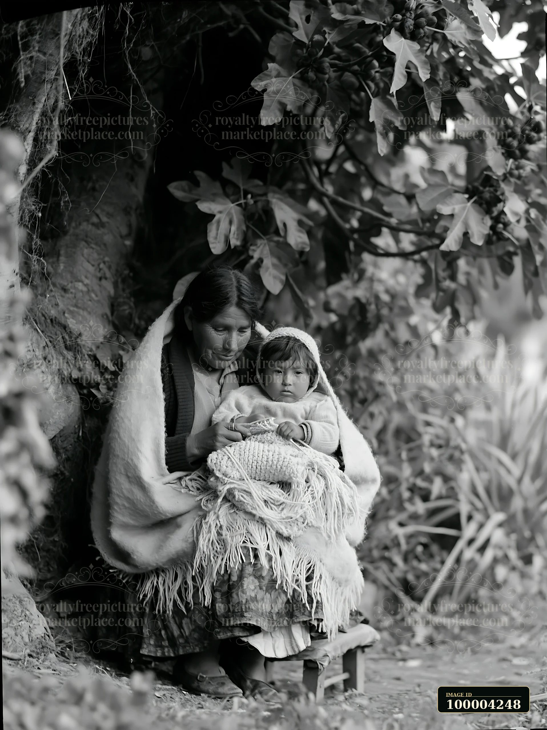 Mother and child sit wrapped in warm blankets beneath a leafy tree in a woodland setting.