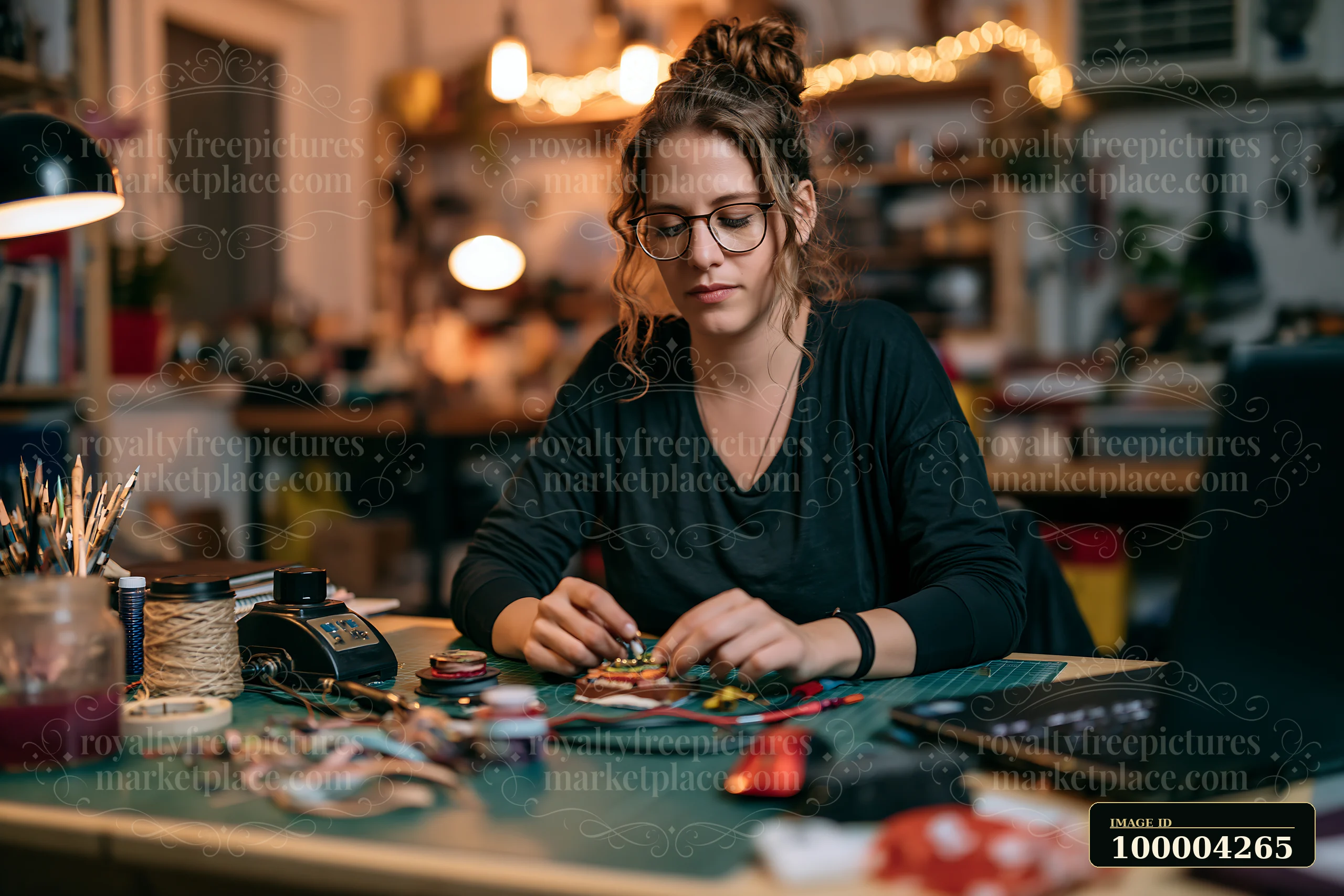 Woman wearing glasses crafts jewelry at a cluttered desk in a warmly lit home workspace.