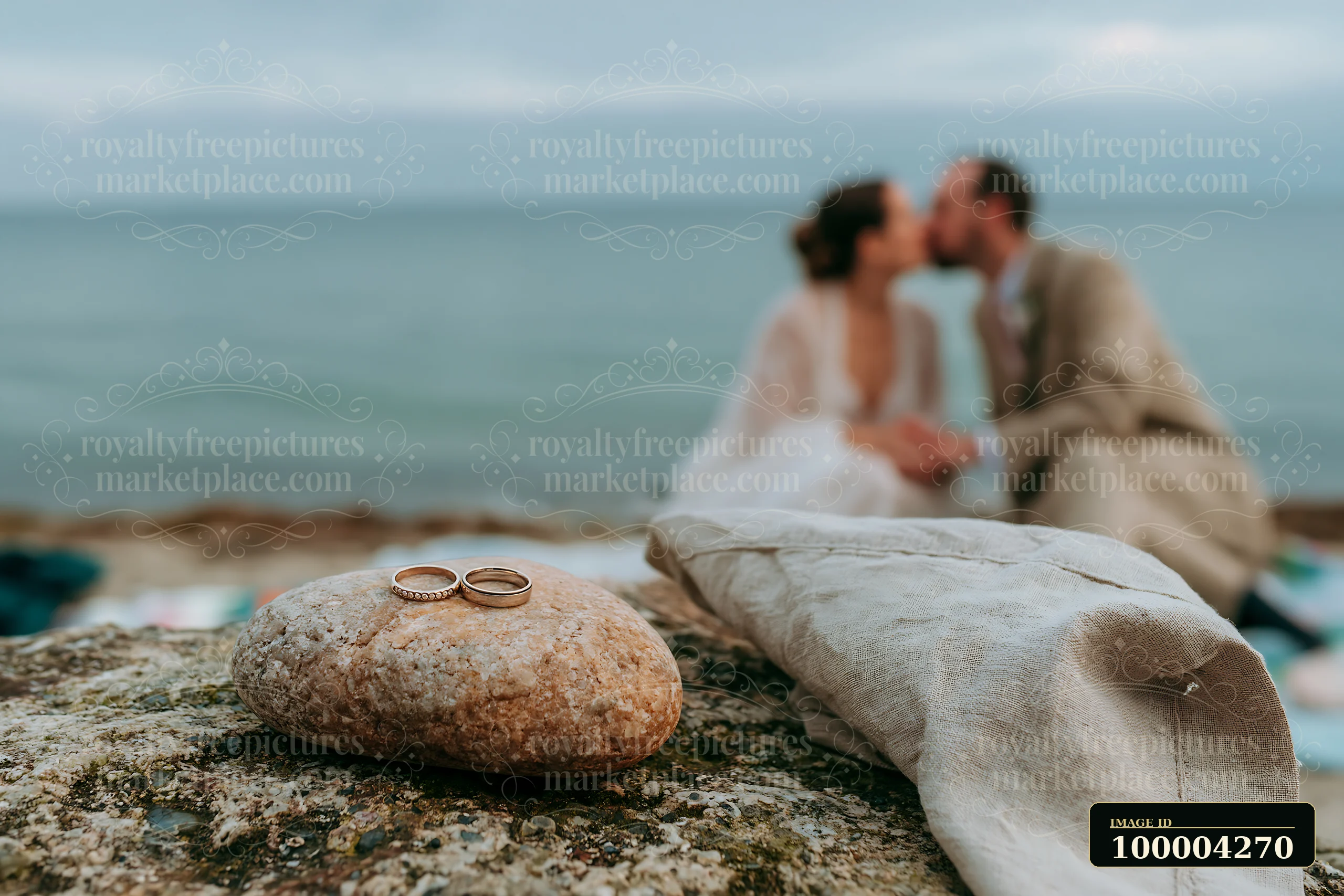 A couple kisses softly while wedding rings rest on a stone nearby.