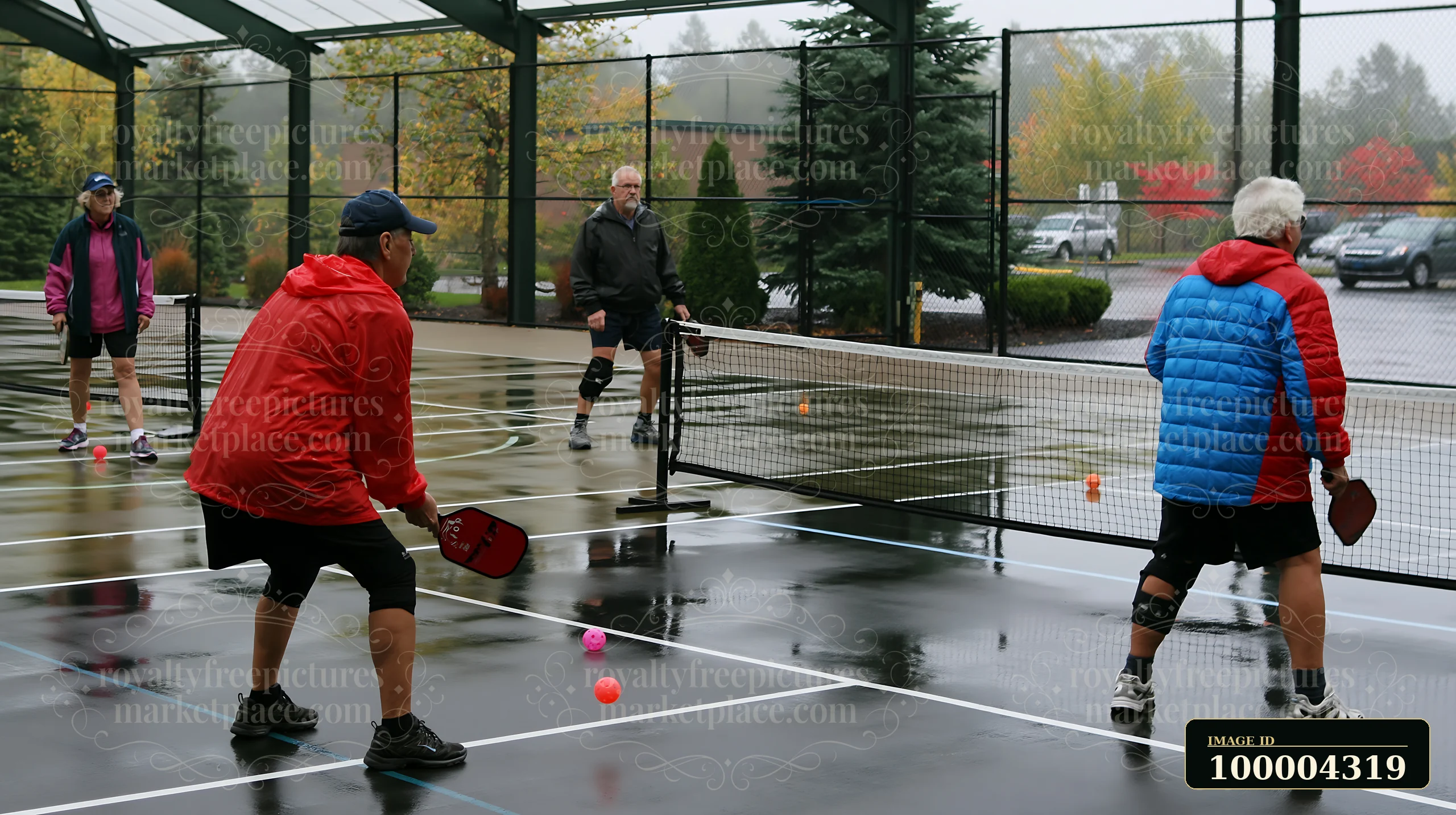 Four older adults play pickleball with paddles on a rain-soaked outdoor recreational court.