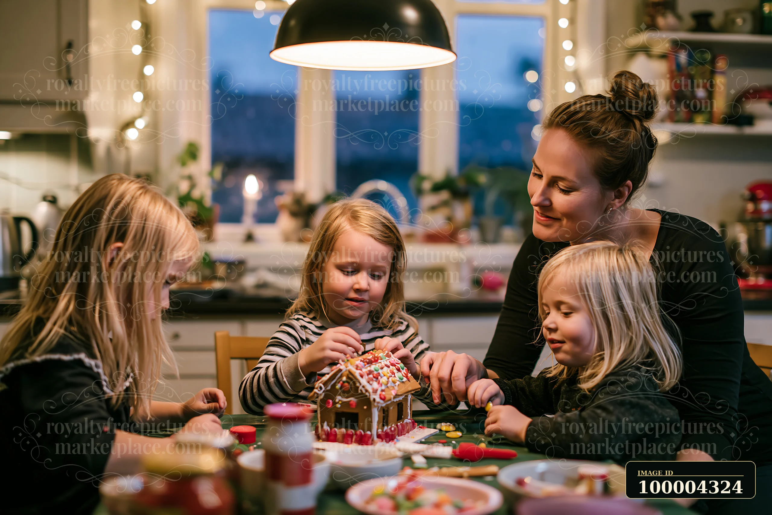 A mother and three children decorate a gingerbread house with icing and candy at a kitchen table.