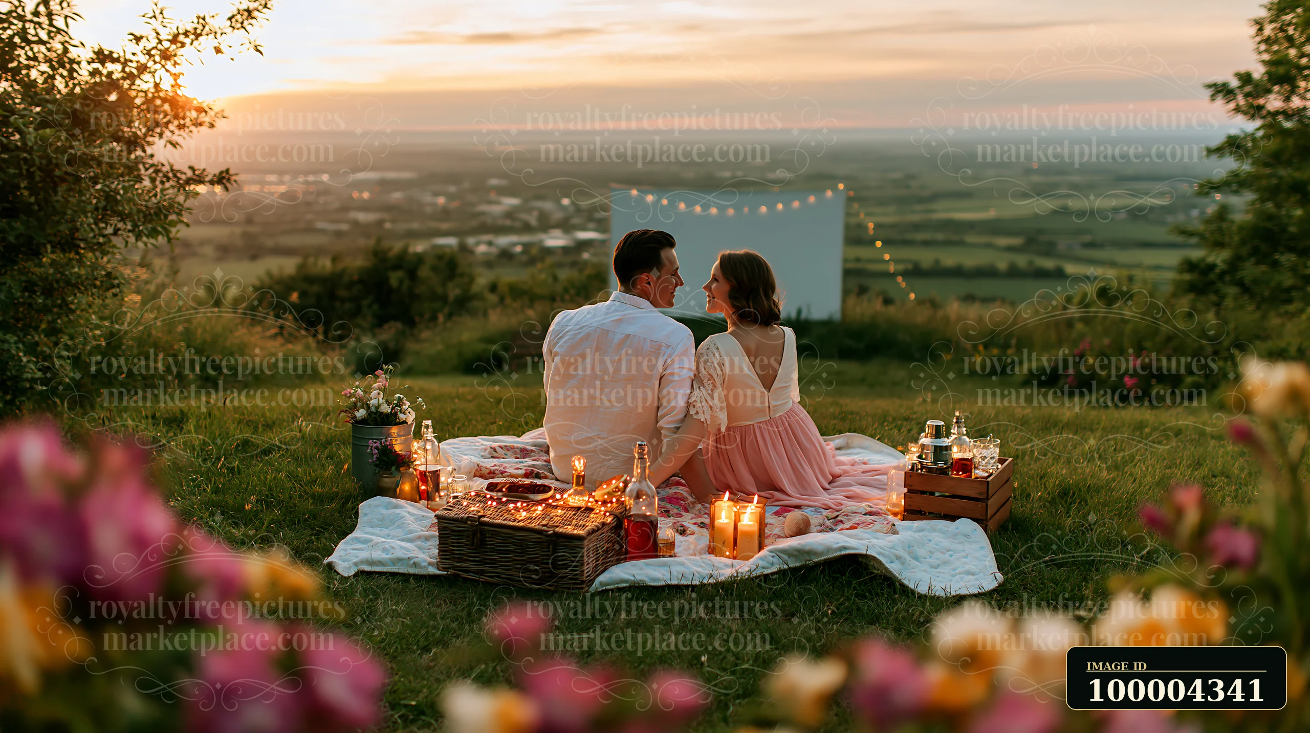 A couple sits together on a picnic blanket surrounded by flowers, enjoying a setup for outdoor cinema as the sun sets over a scenic landscape.