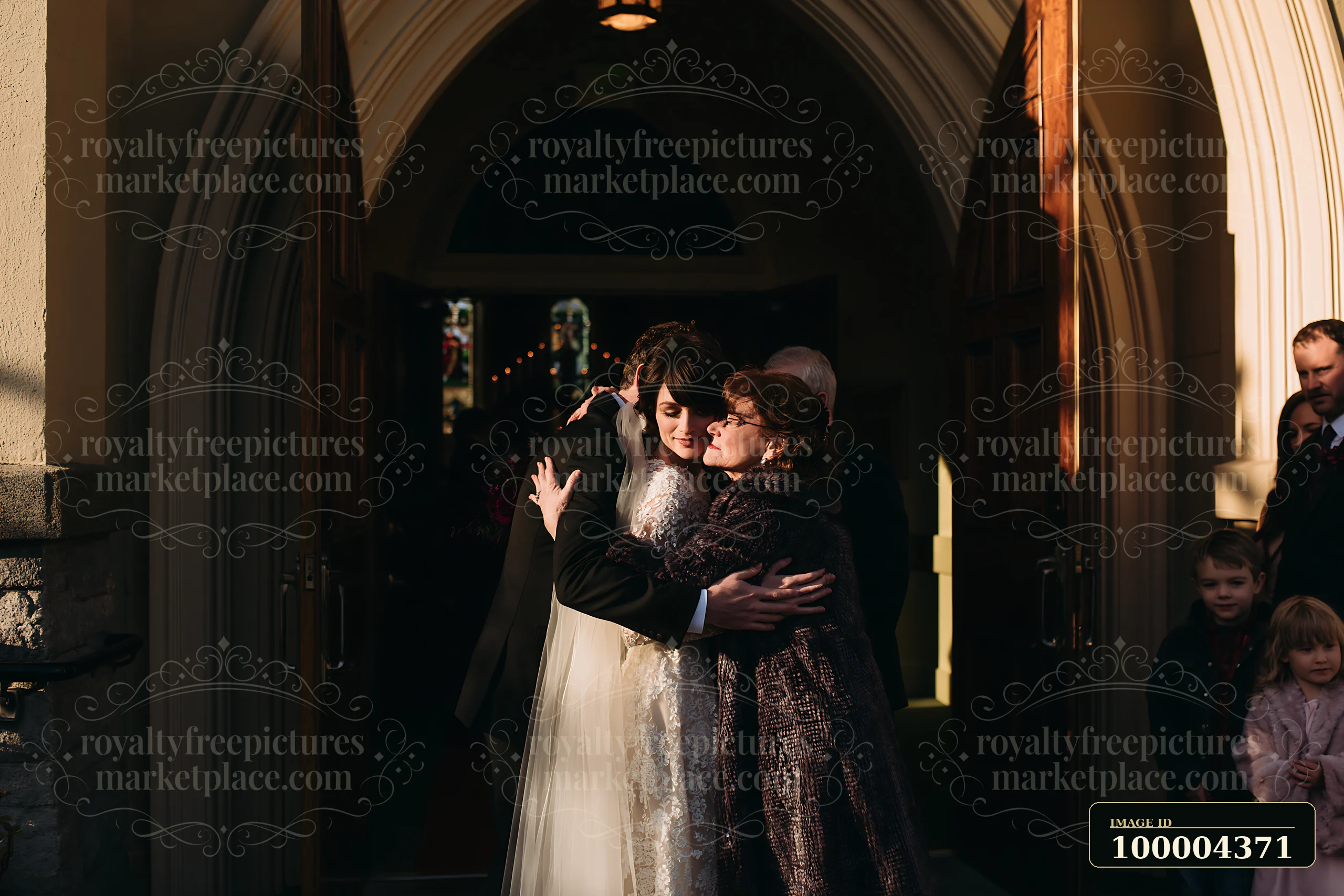 Newlywed couple embraces family beneath a sunlit church doorway arch while wedding guests gather nearby.
