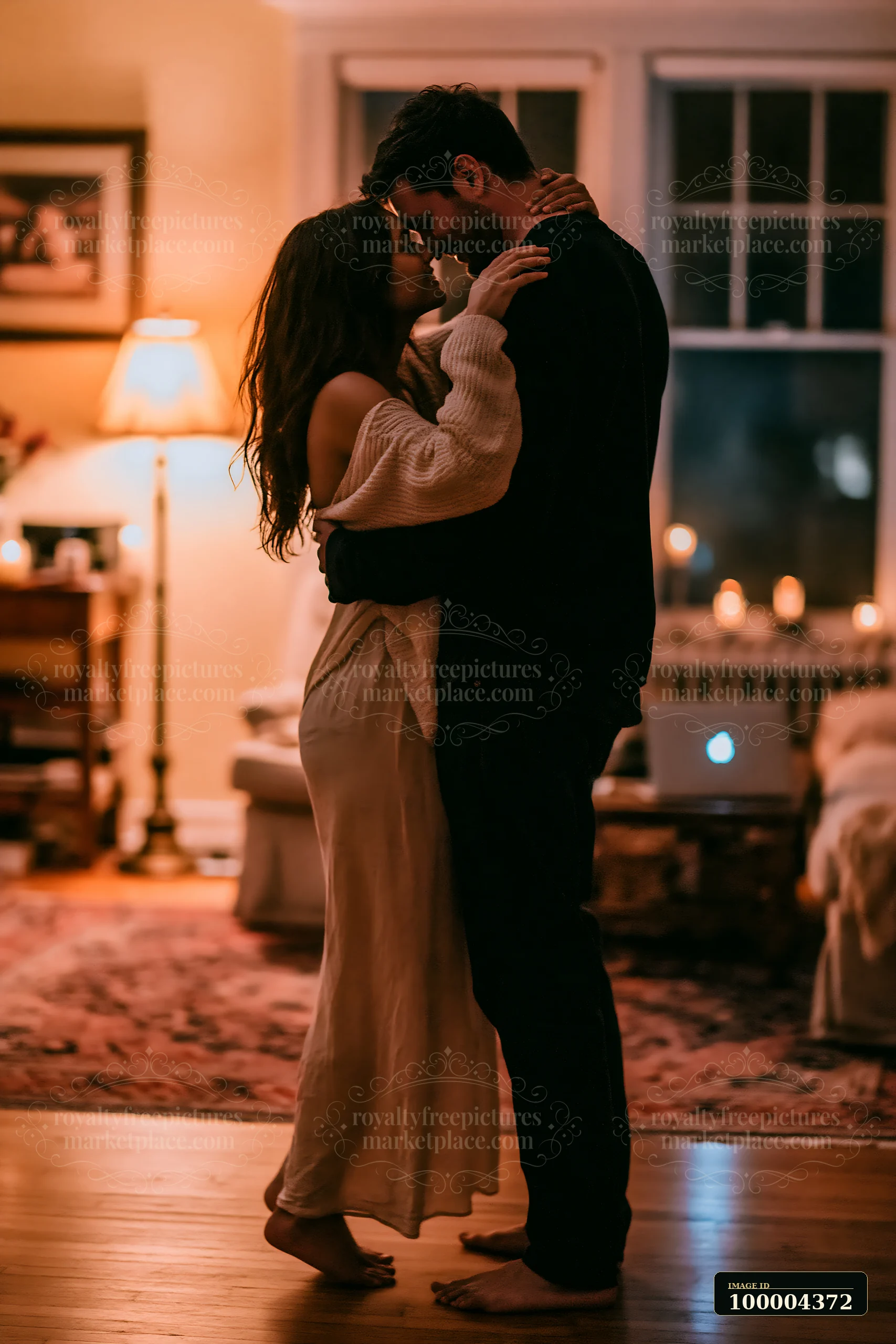 Couple embraces and dances barefoot on a wooden floor in a warmly lit living room.
