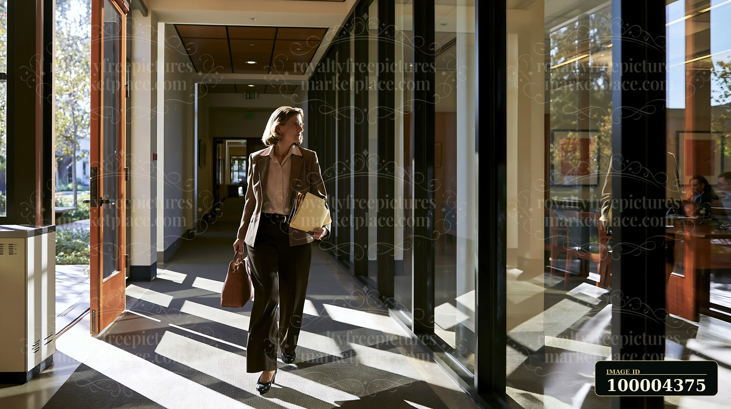 An executive woman walks through a modern office hallway, carrying work documents and a handbag.
