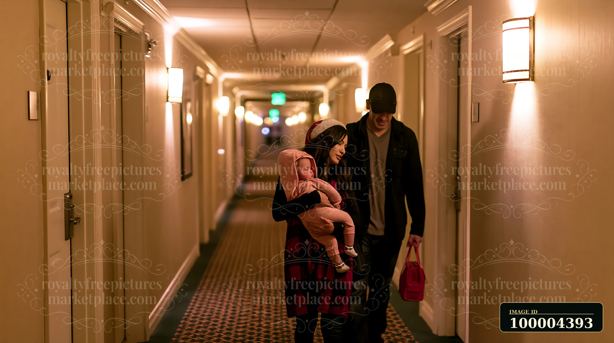 Mother carries a sleeping child while the father walks beside her through a warmly lit hotel hallway.
