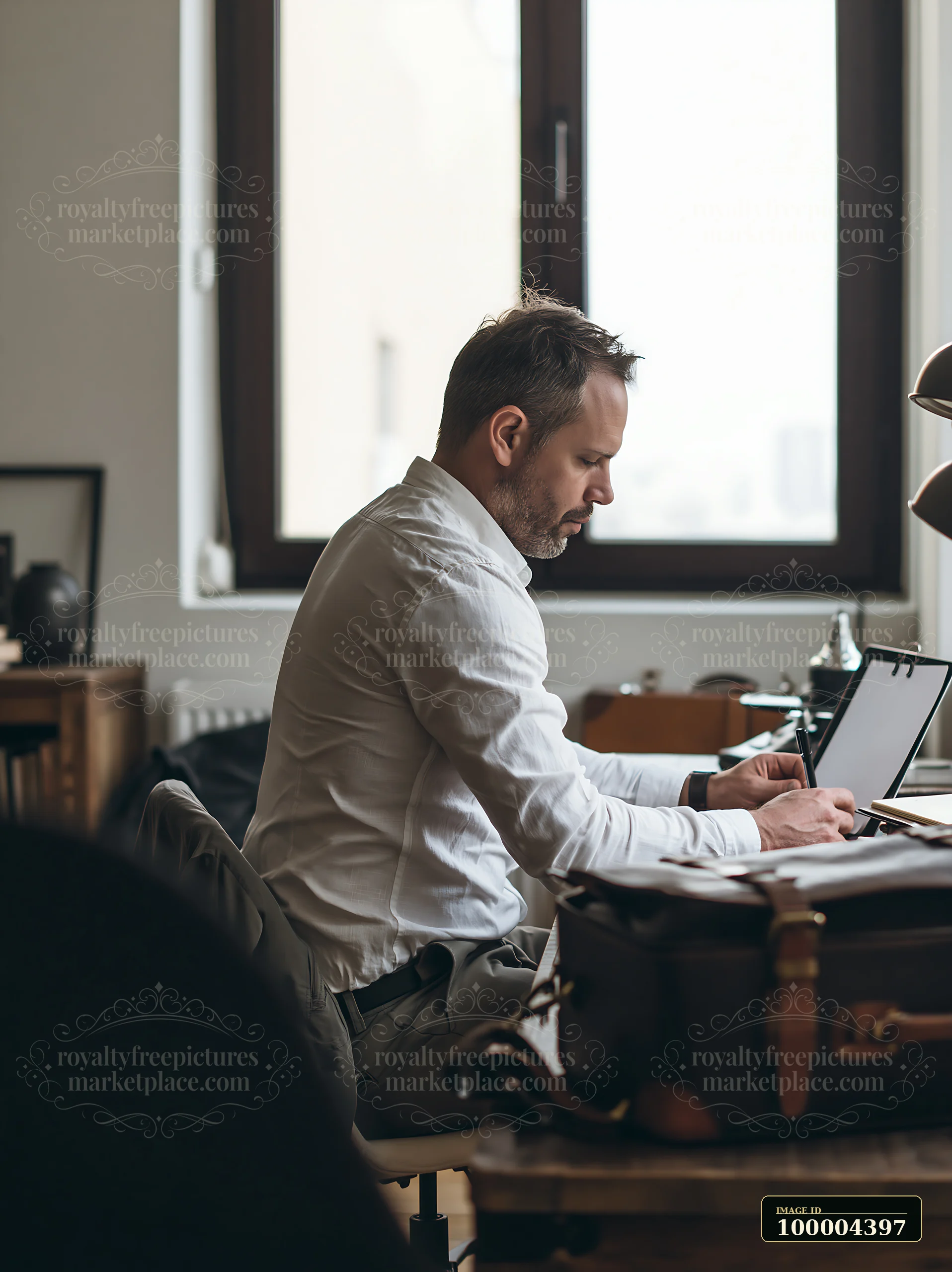 Businessman writes in a notebook at a desk beside a bright window in a home office.