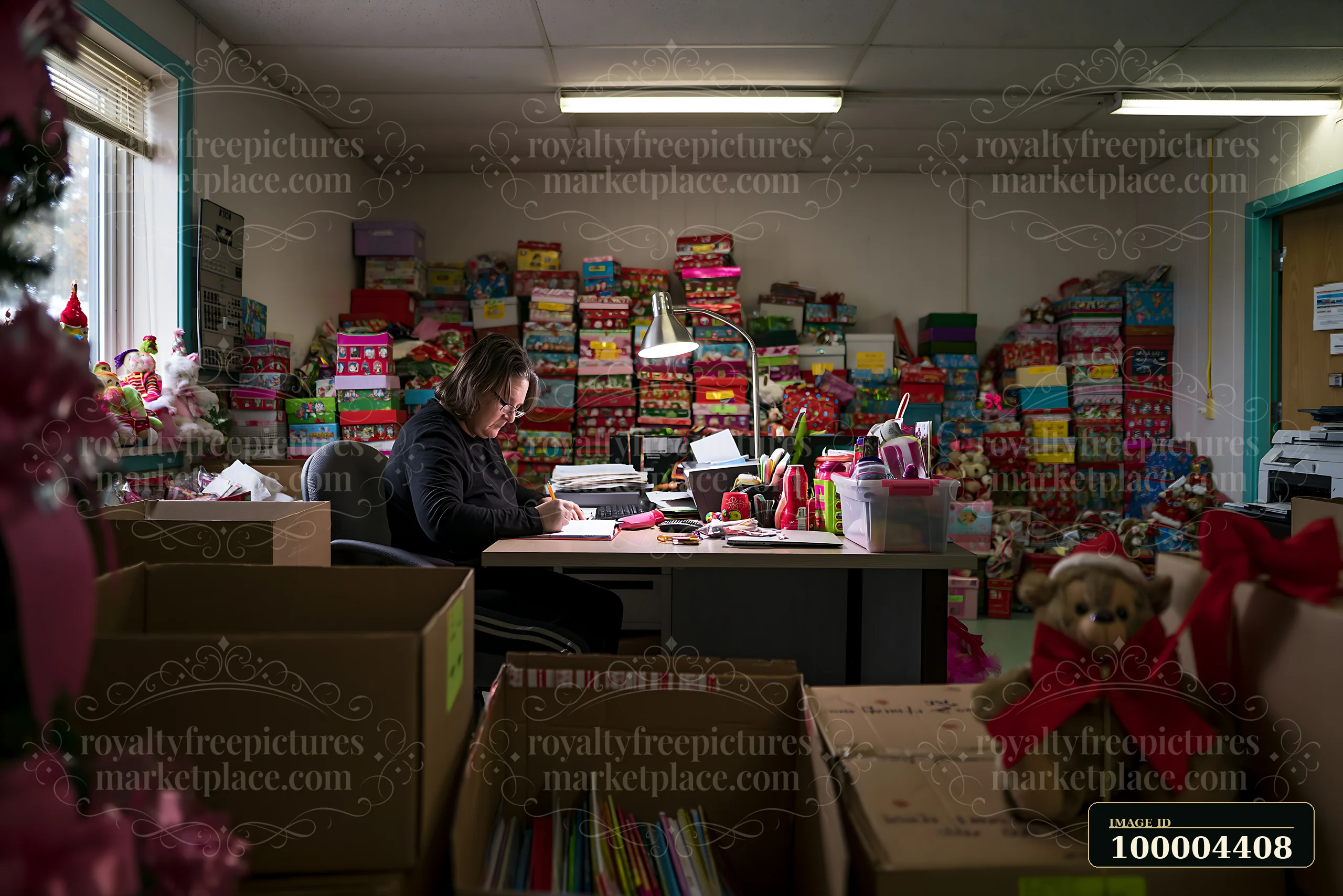 Person writes at a desk surrounded by colorful gift boxes, toys, supplies, and cardboard boxes.
