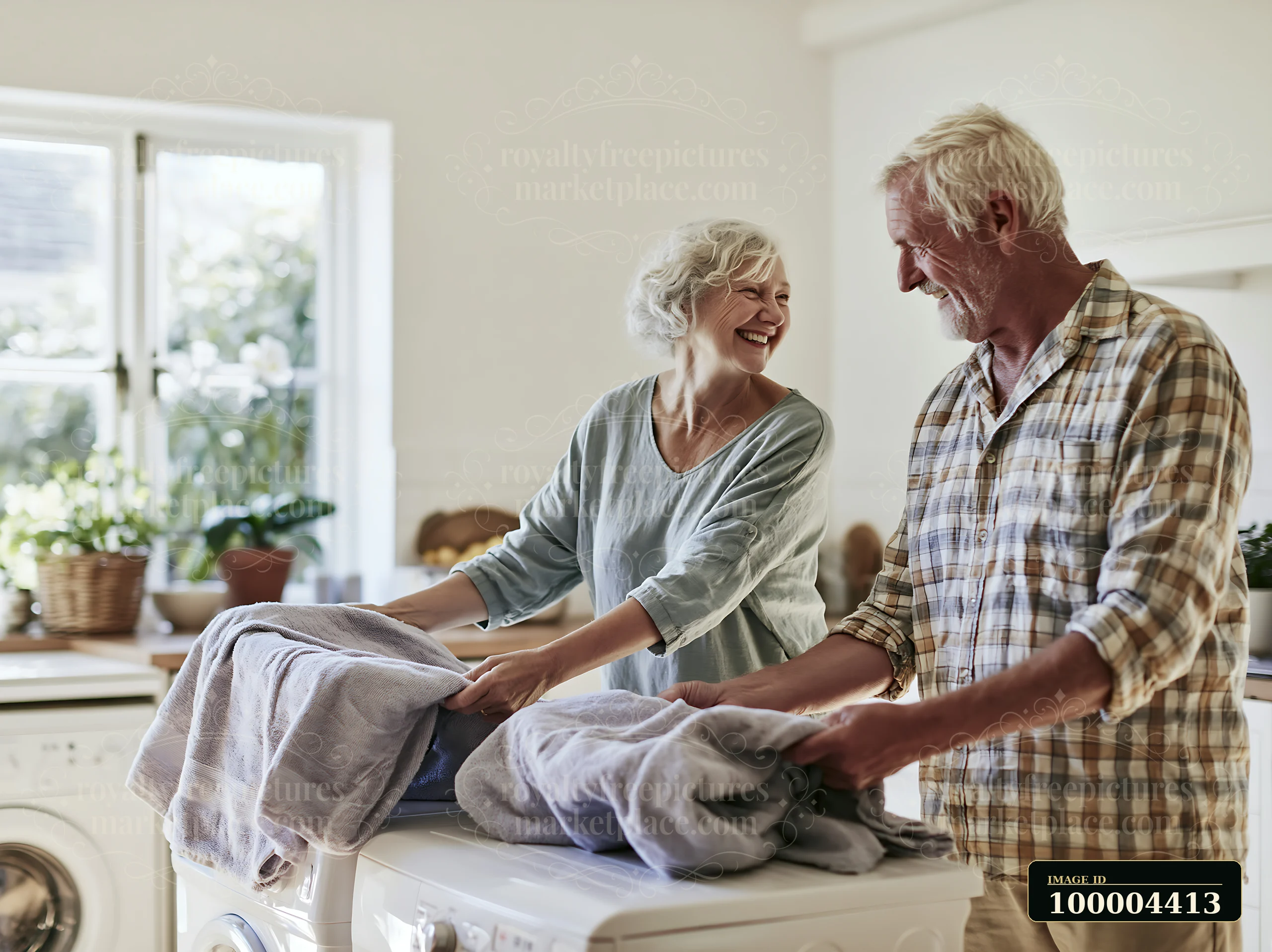 Elderly couple smiles at each other while folding towels together in a bright home laundry area.