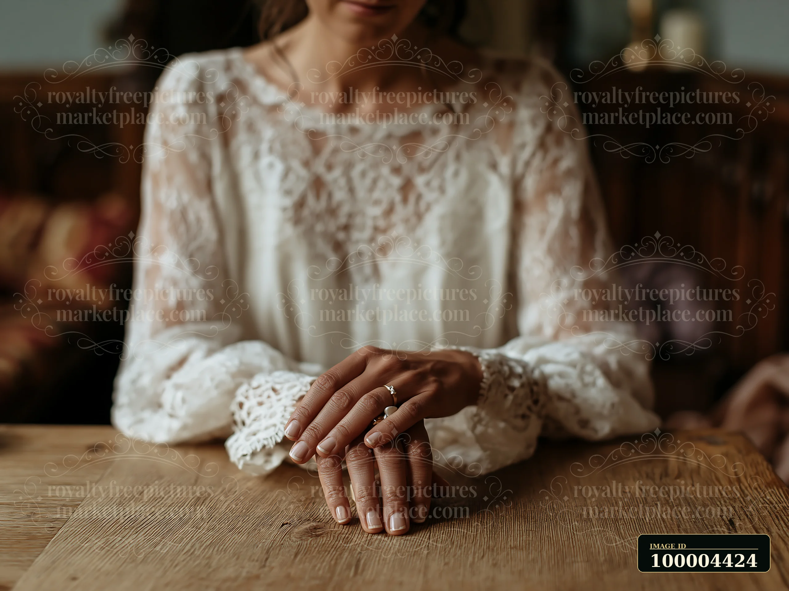 Bride in a lace wedding dress rests her hands on a wooden table in warm interior light.