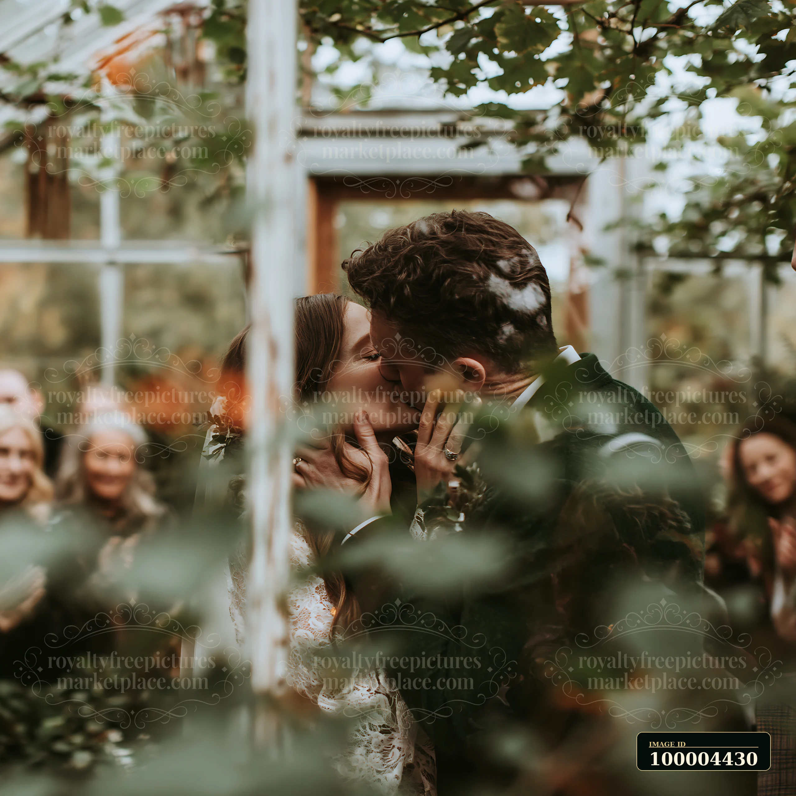 Bride and groom kiss in a leafy outdoor wedding setting while guests watch in the blurred background.