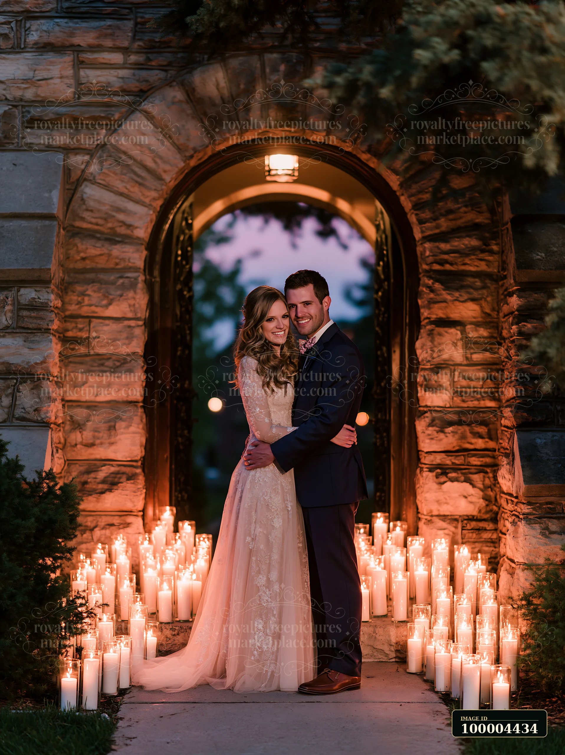 Newlywed couple embraces beneath a candlelit stone church doorway arch surrounded by evergreen branches.