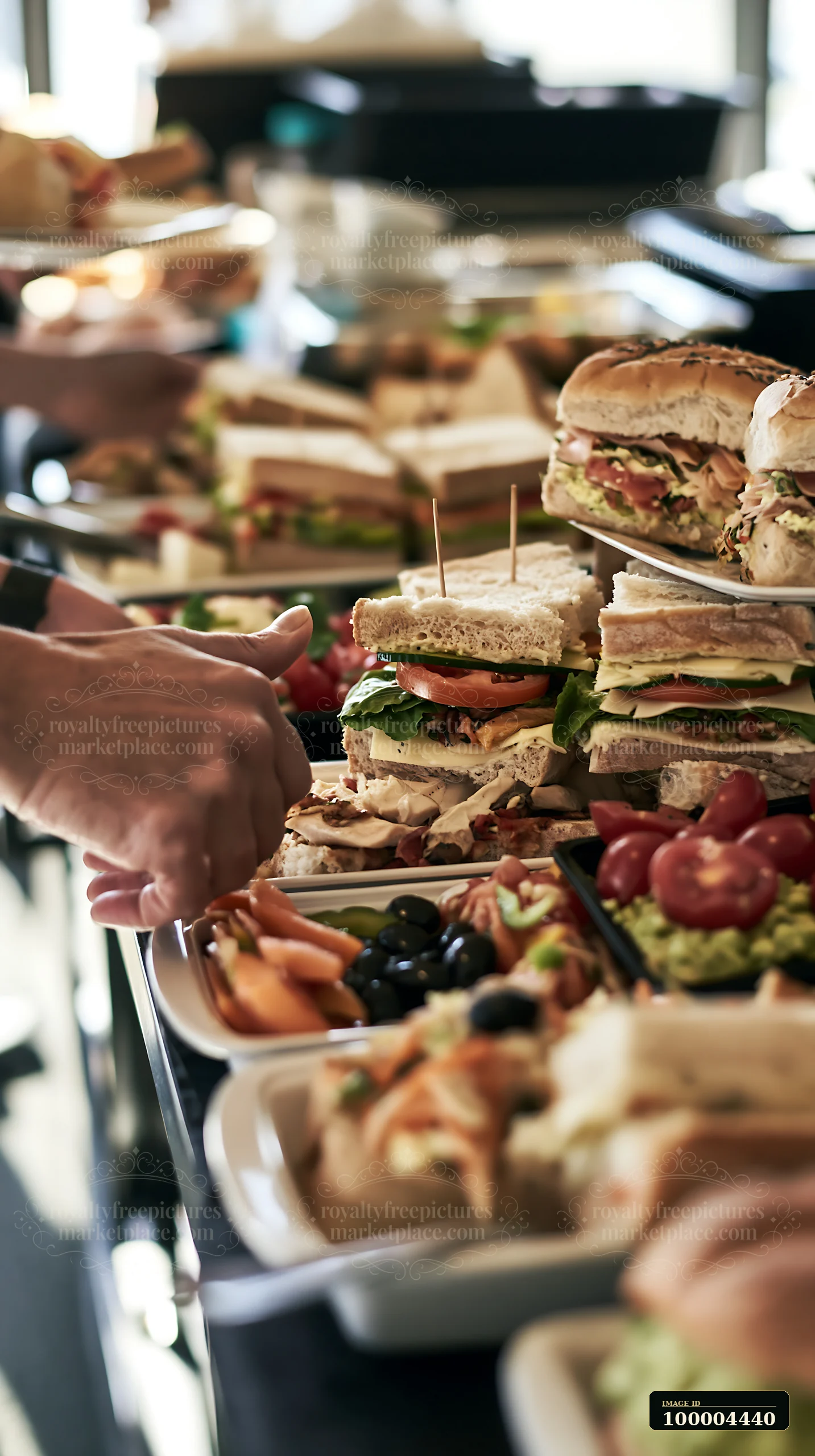 Stacked sandwiches and vegetables fill a workplace lunch buffet as a hand selects food.