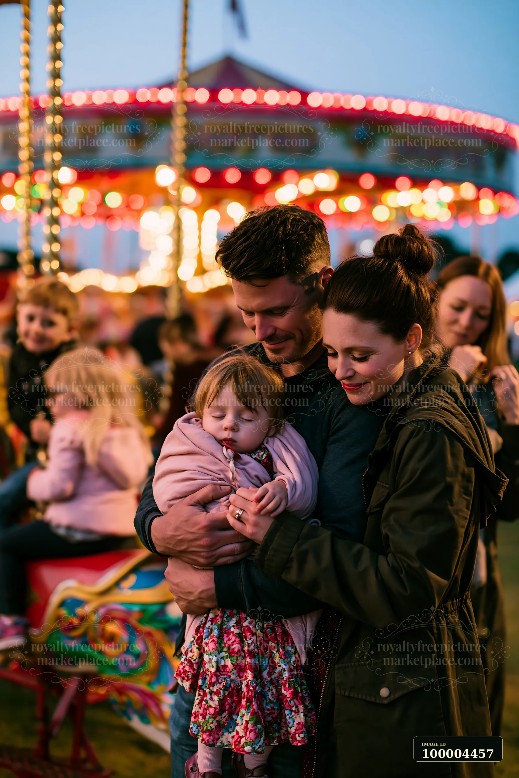 Parents hold a sleeping child beside a brightly lit carousel at dusk.