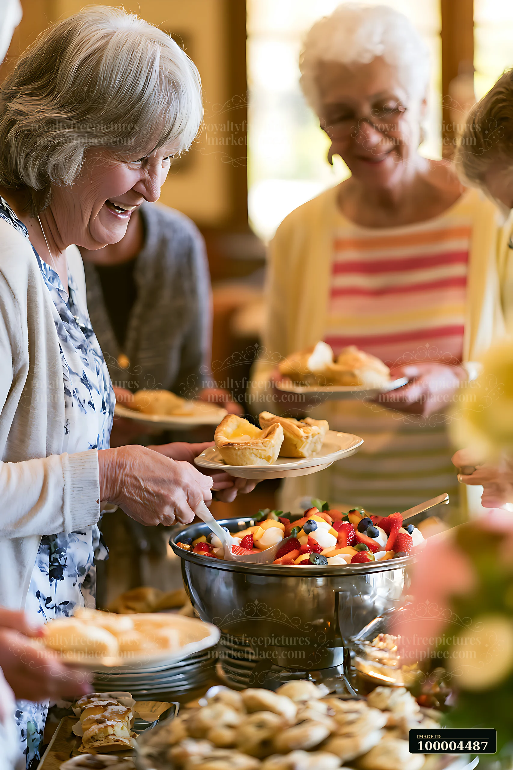 Older adults serve pastries beside a fruit salad buffet at a warmly lit indoor celebration.