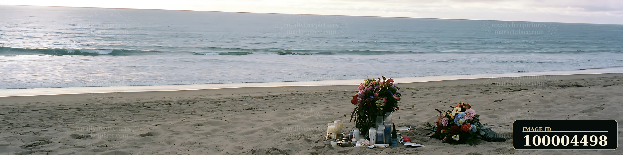 Flowers and candles form a memorial on a sandy beach beside calm ocean waves.