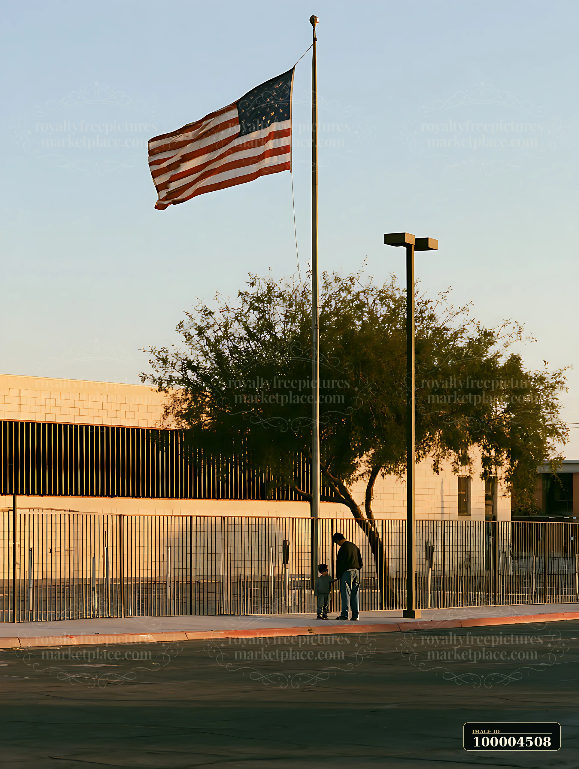 American flag waving beside a fenced school building with two people walking on the sidewalk.