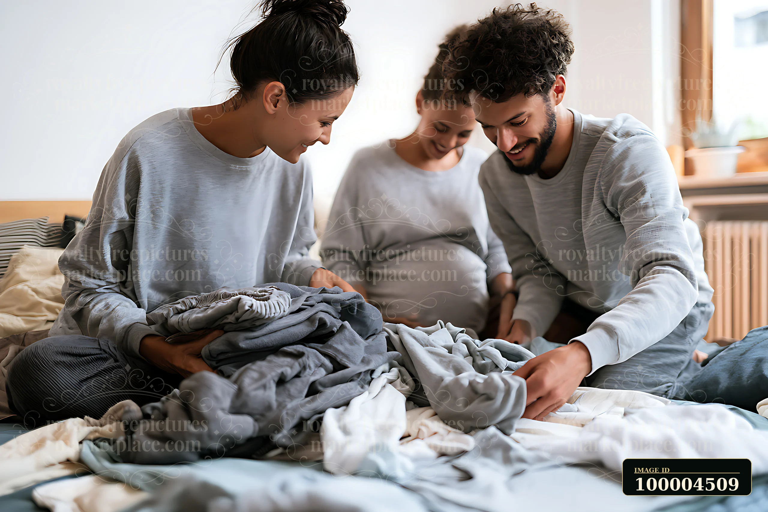 Pregnant couple folds baby clothes together on a bed at home while another woman looks on.