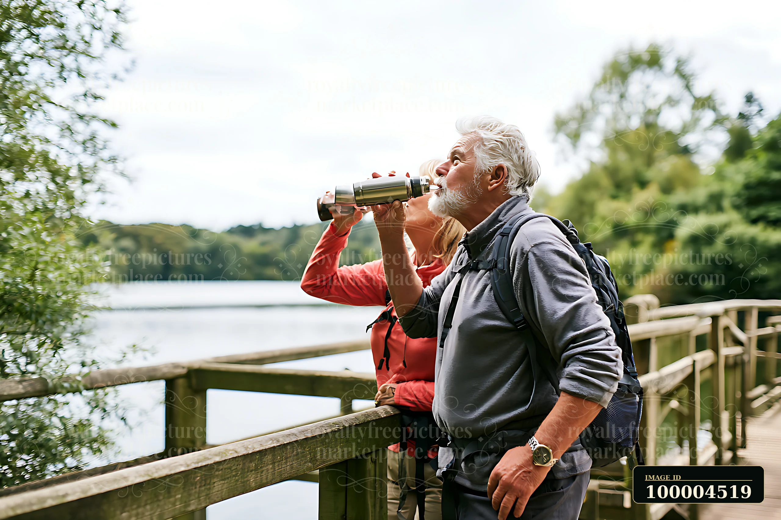 Older couple with backpacks stands on a wooden footbridge beside a lake, looking through binoculars.