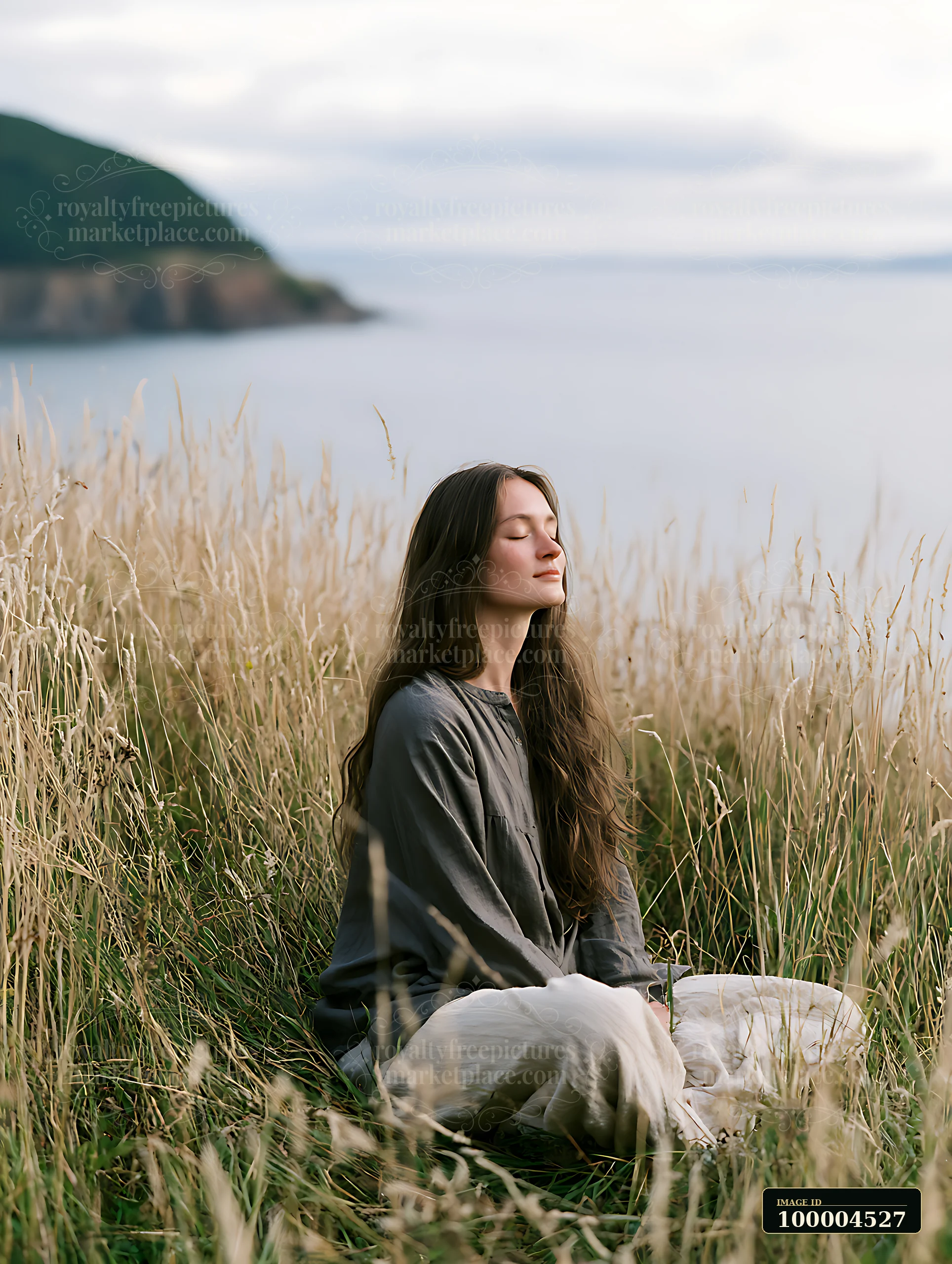 Woman with closed eyes sits in tall coastal grass beside calm water and a distant headland.