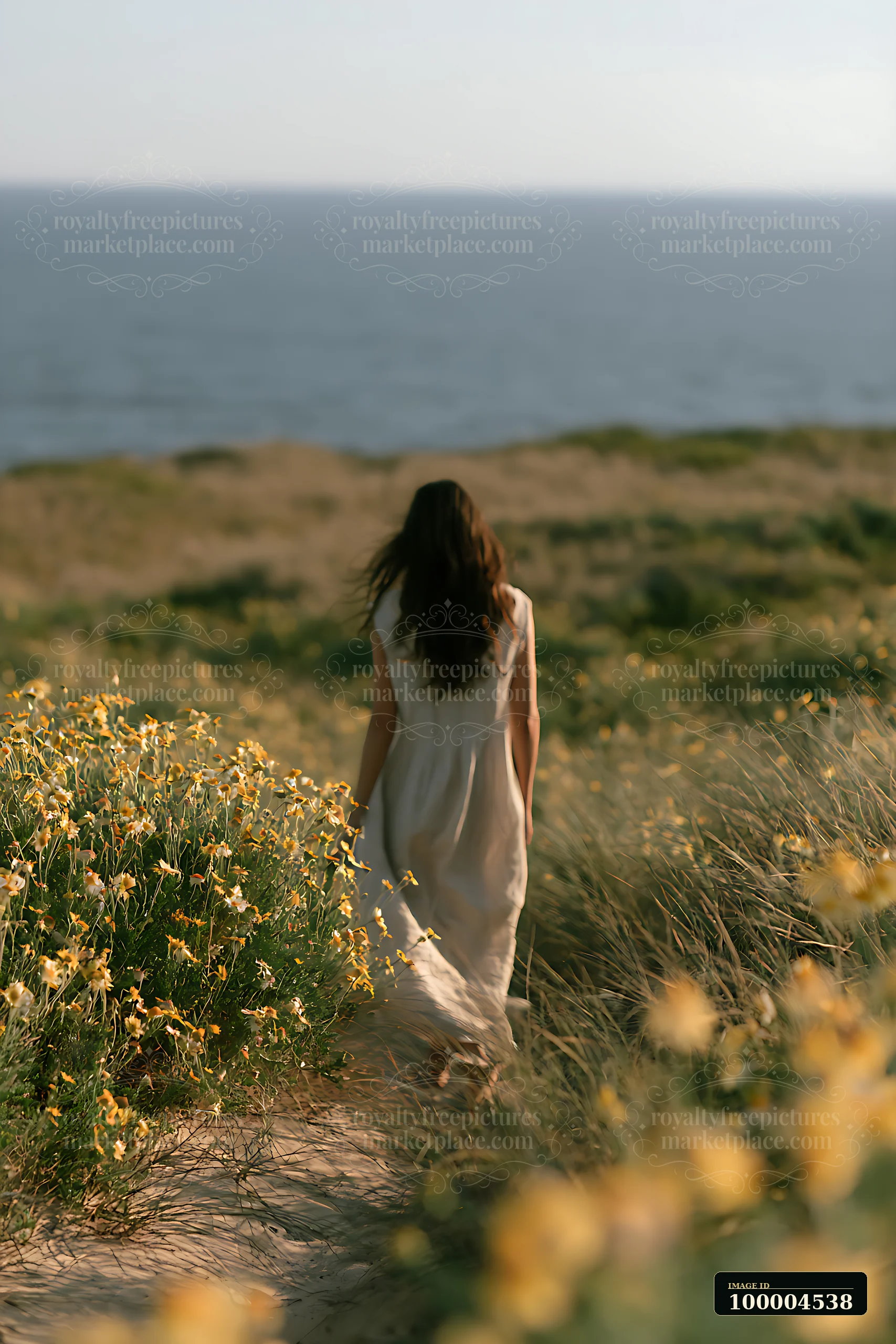 A woman walks along a sandy path lined with bright flowers, leading towards a wide ocean view under clear skies.