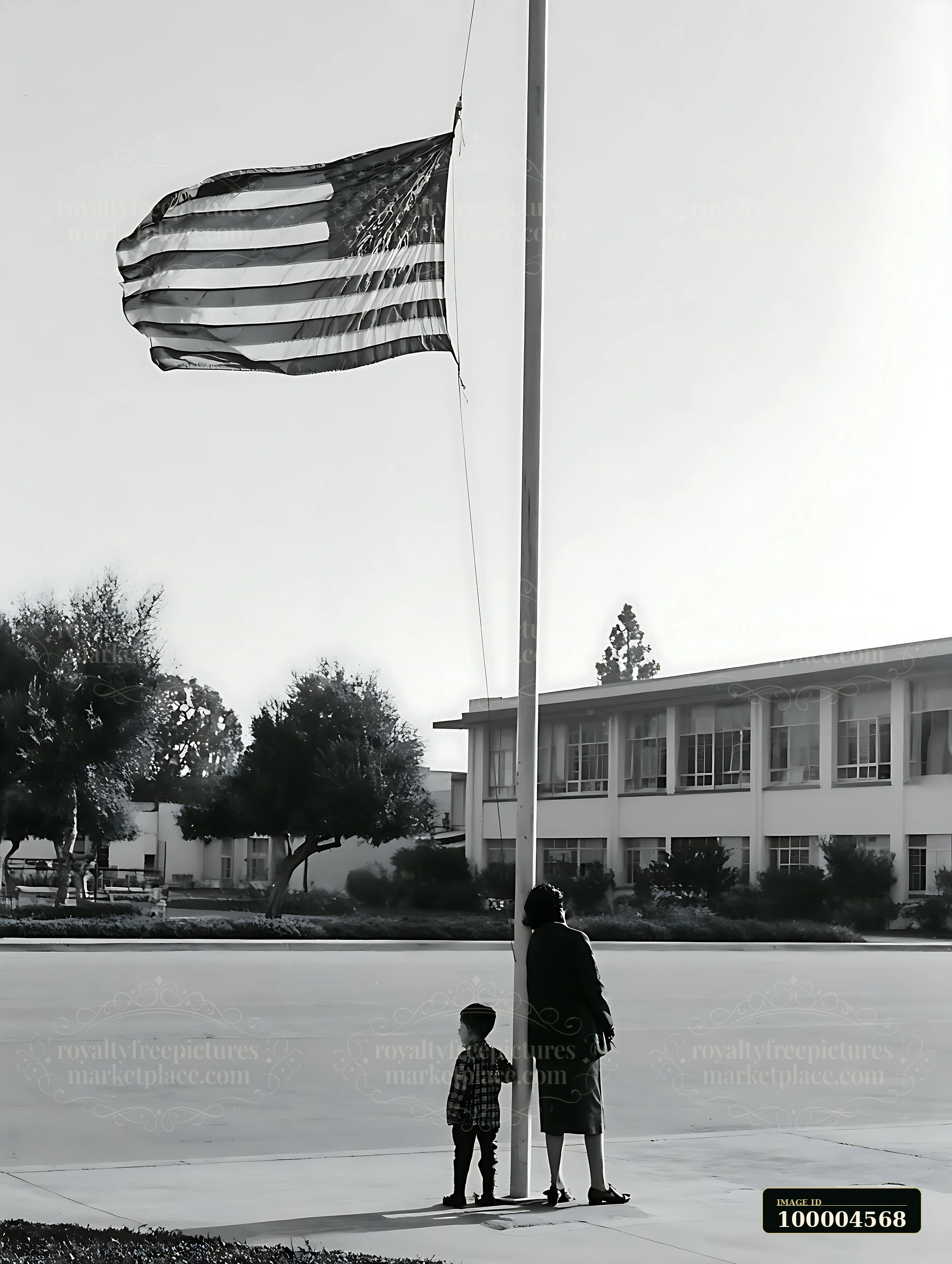 A child and an adult stand together under an American flag waving beside a school building.