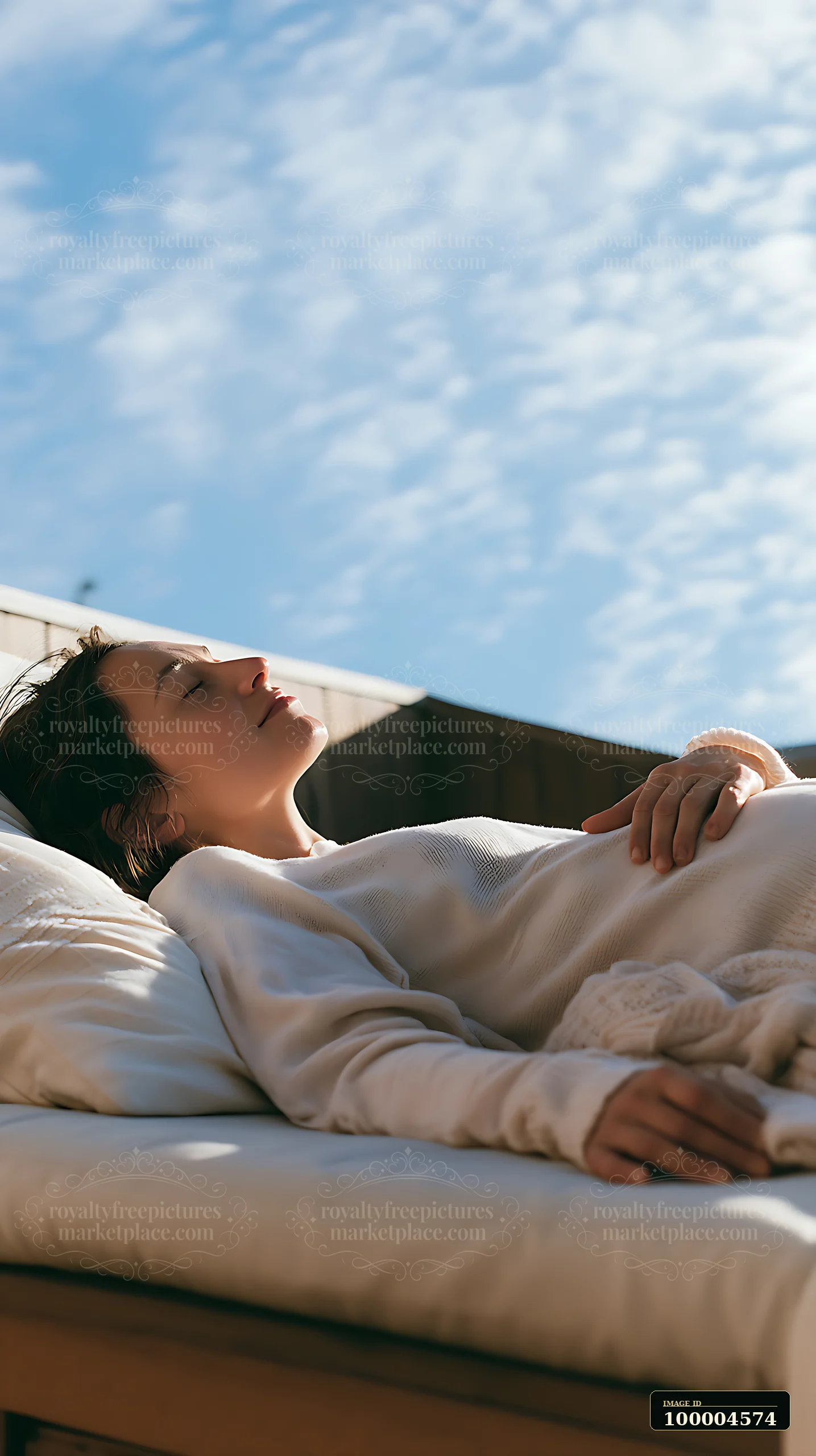 Pregnant woman rests on a bed with one hand on her belly beneath soft natural light and a blue sky.
