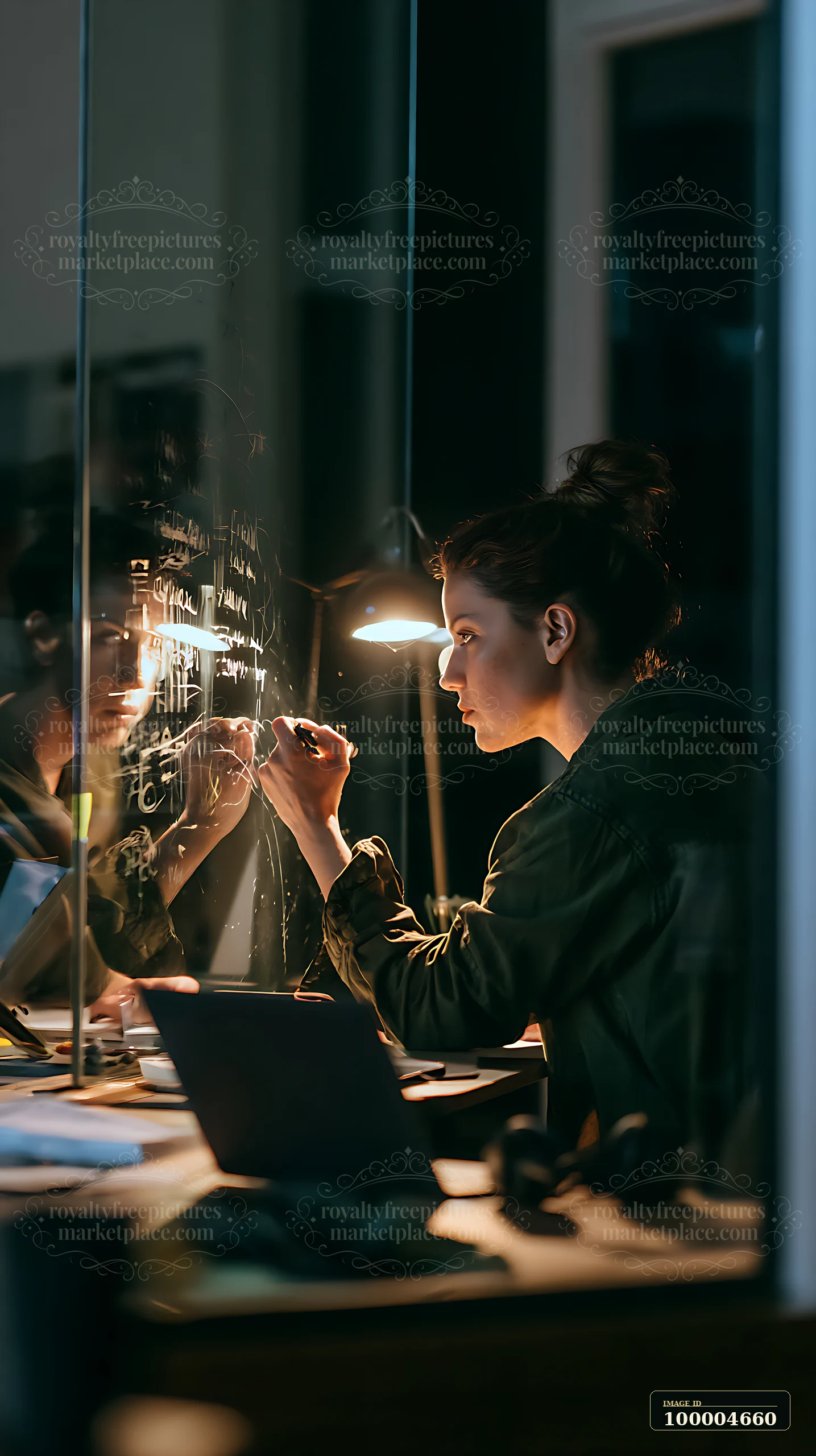 Businesswoman writes notes on a glass board in a dimly lit office beside a laptop.