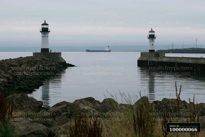 Twin harbor lighthouses frame a cargo ship sailing across calm water beneath an overcast sky.