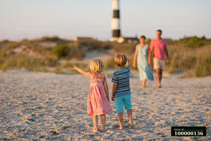 Two children walk across a sandy coastal path toward a lighthouse while two adults follow behind.