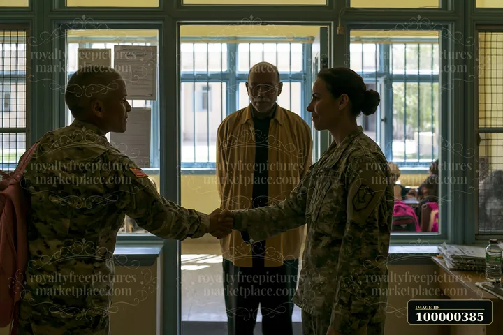 Two soldiers greet each other with a handshake in a room, while an observer watches.