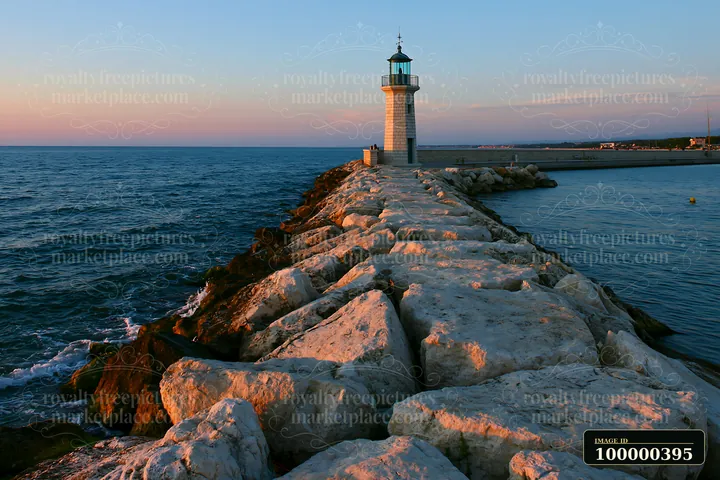 A lighthouse stands on a rocky jetty as the sun sets over the ocean.