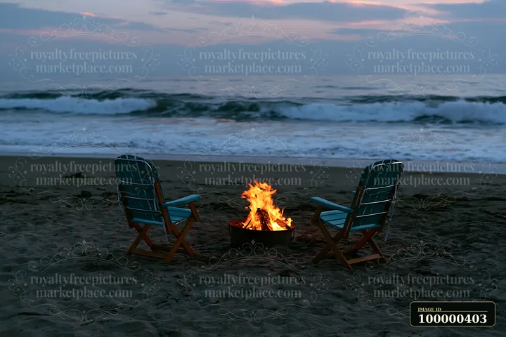 The scene shows two chairs facing a bonfire on the sand at a coastline during sunset.