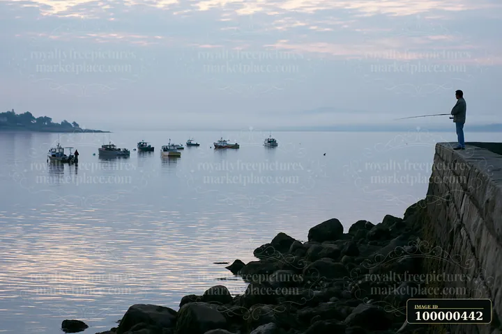 A person stands on a protective sea wall along the coastline, casting a fishing line into calm water while several fishing boats are anchored nearby.