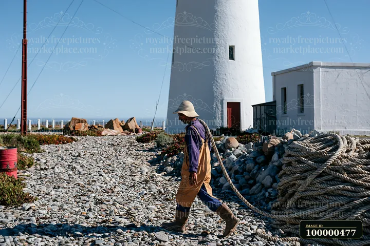 A lighthouse maintenance worker at work walks along a rocky path near the lighthouse, carrying ropes and preparing for tasks to ensure the structure is function