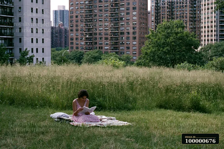 A woman sits on a blanket in a green urban meadow among buildings, reading a book.