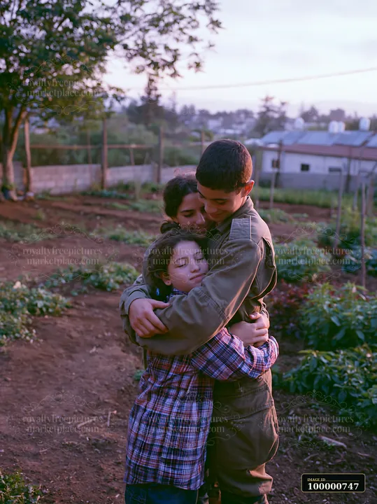 Two children hug a third child in a garden surrounded by plants.