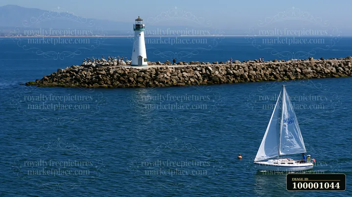 A lighthouse stands on a rocky pier while a sailboat moves through the water nearby.