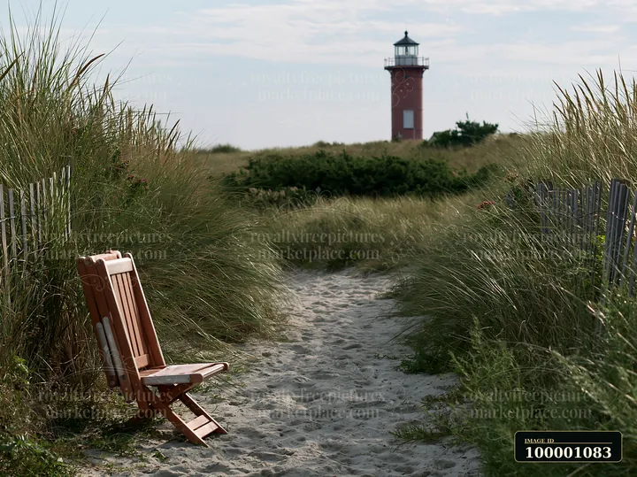 A wooden chair sits on a sandy path leading to a lighthouse framed by coastal dunes.