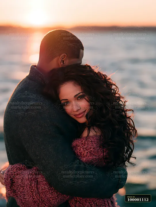 A couple hugs closely near the water as the sun sets on the horizon.