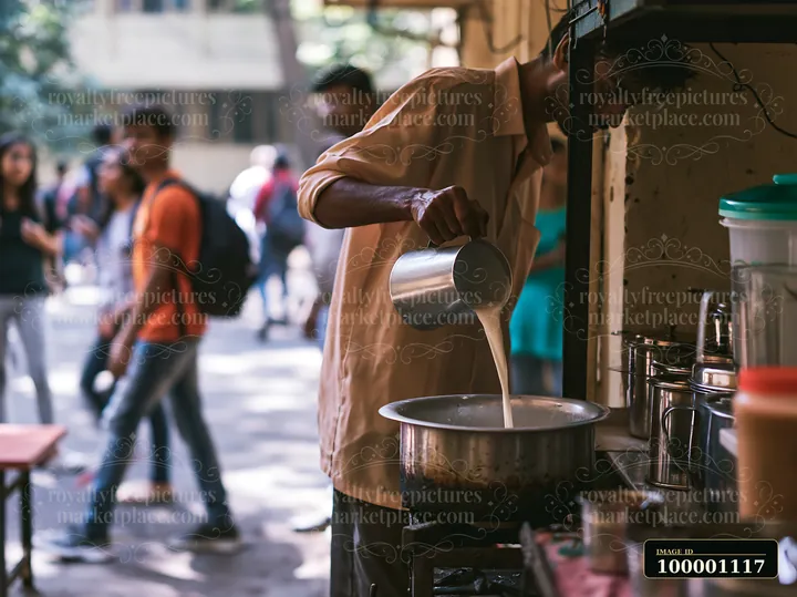 A street food vendor is pouring milk into a pot of tea at a bustling outdoor setting with people walking by.