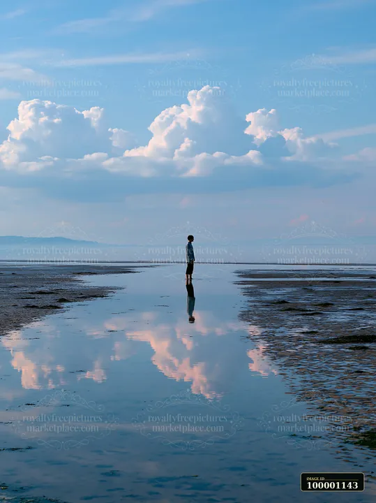 A lone person stands in shallow water at the coastline, surrounded by cloud reflections and clear skies.